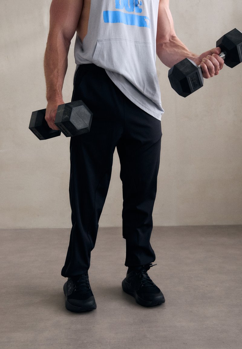 Hoodie cinza sem mangas, calças de treino pretas e sapatilhas pretas. Pesos em cada mão, exibindo um design hexagonal e um agarre texturizado.