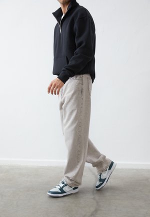 Sudadera de fleece negra con medio cierre y bolsillo frontal, combinada con pantalones sueltos de corte ligero en gris claro y zapatillas verdes y blancas. Textura suave, ajuste casual.