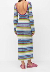 Robe maxi en tricot avec un dos échancré profond, présentant des rayures horizontales multicolores en bleu, vert, violet et blanc. Complétée par des sandales roses.