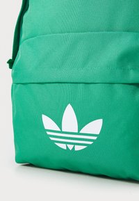 Πράσινο σακίδιο από ύφασμα με υφή και λευκό λογότυπο με τρεις ρίγες. Διαθέτει μπροστινή τσέπη και ρυθμιζόμενους ιμάντες ώμου.