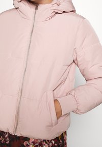 Růžová péřová bunda s kapucí, vybavená předním zipem, elastickými manžetami a bočními kapsami. Vyrobena z hladkého, lehkého materiálu.