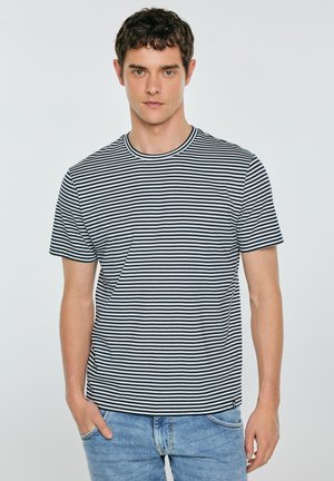 Mężczyzna stoi, ubrany w czarno-białą koszulkę w poziome paski i jasnoniebieskie dżinsy, ręce rozluźnione, zwrócony twarzą do przodu na jednolitym tle.