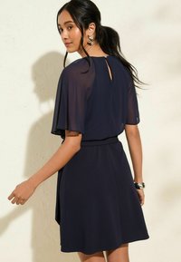 Robe navy à manches courtes en tissu chiffon, silhouette fluide et détail de dos en forme de clé. Présente une taille cintrée et une jupe légèrement évasée.