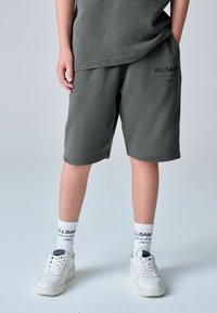 Osoba oblečená v sivých textúrovaných šortkách a zodpovedajúcom topu, s bielymi ponožkami a bielymi teniskami, stojaca na svetlom pozadí.