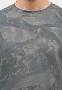 Κοντινή λήψη ενός ατόμου που φοράει γκρι T-shirt με σχέδιο παραλλαγής, αφηρημένα σχήματα και απαλούς τόνους.