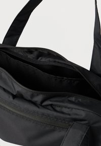 Črna torba iz najlona z zadrgo na vrhu in notranjim žepom. Ima trpežne naramnice in gladko teksturo, zasnovano za trajnost.