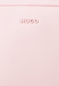 Ružový faux kožený povrch s embosovaným logom "HUGO", hladká textúra a jemný zrnitý vzor. Kompaktný obdĺžnikový dizajn.