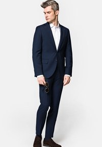 Granatowy garnitur dopasowany z pasującymi spodniami, biała koszula i brązowe buty. Model trzyma okulary przeciwsłoneczne, prezentując elegancki, nowoczesny design.