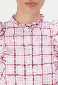 Femme portant un chemisier à carreaux rose et blanc avec col et manches à volants, boutonné à l'avant.