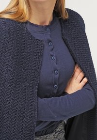 Marinblå texturerad cardigan över en marinblå långärmad skjorta med knappar. Fyra knappar syns, med ribbade muddar, och en arm korsad framför kroppen.