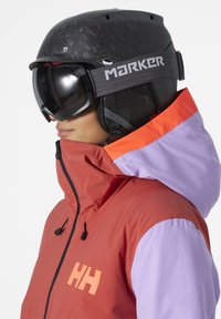 Skijakke i røde og lavendeltoner, med høy krage, svarte glidelåser og en logo på brystet. Akkompagnert av en svart skihjelm og goggles.