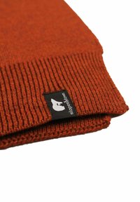 Berretto lavorato a maglia in arancione ruggine con un motivo a coste, con un'etichetta nera con logo bianco cucita sul bordo. Texture morbida, design piegato.