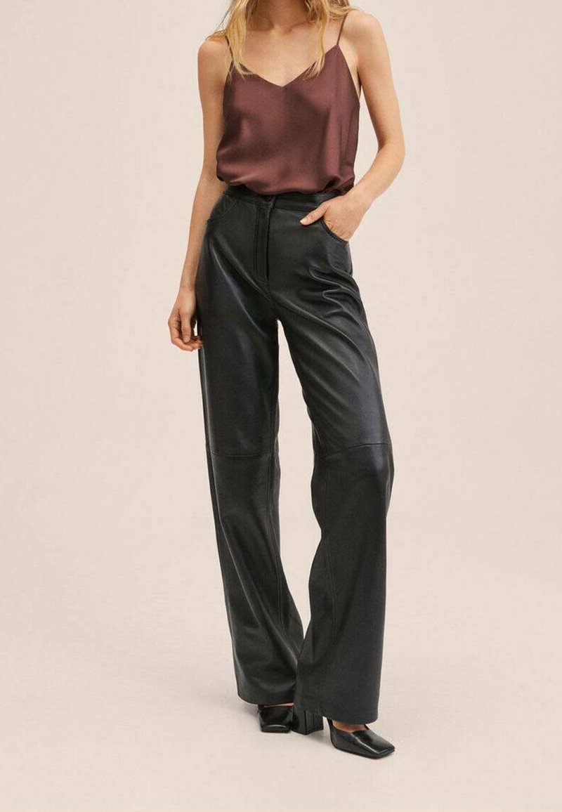 Femme portant un haut marron sans manches avec un pantalon noir taille haute à jambes larges et des chaussures noires à bout carré, debout avec une main dans la poche.
