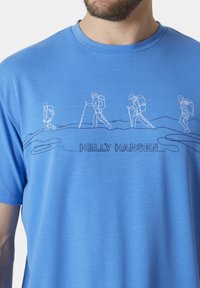 Camiseta azul con un gráfico de senderismo en blanco, que representa a cuatro excursionistas con mochilas y bastones. El texto dice "HELLY HANSEN."