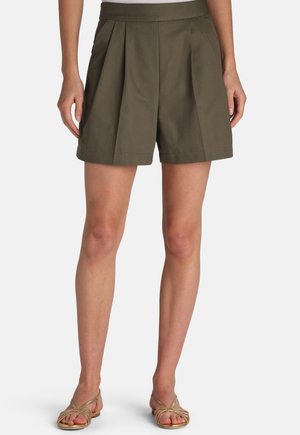 Žena v olivovo zelených plisovaných šortkách s vysokým pásom a zlatých plochých sandáloch s remienkami, stojaca na jednoduchom bielom pozadí.