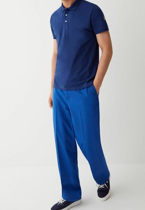 Homme portant un polo bleu foncé à manches courtes, un pantalon ample bleu vif et des baskets bleu foncé avec des semelles et des lacets blancs.