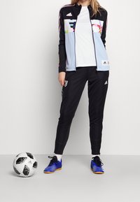 Černá sportovní bunda s světle modrými a barevnými geometrickými vzory, doplněná o černé sportovní kalhoty a modré sportovní boty. Vedle leží černobílý fotbalový míč.