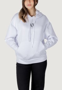Sudadera blanca con capucha, bolsillo frontal, cordón ajustable en la capucha y logo negro en el pecho. El material parece suave, con un corte holgado.