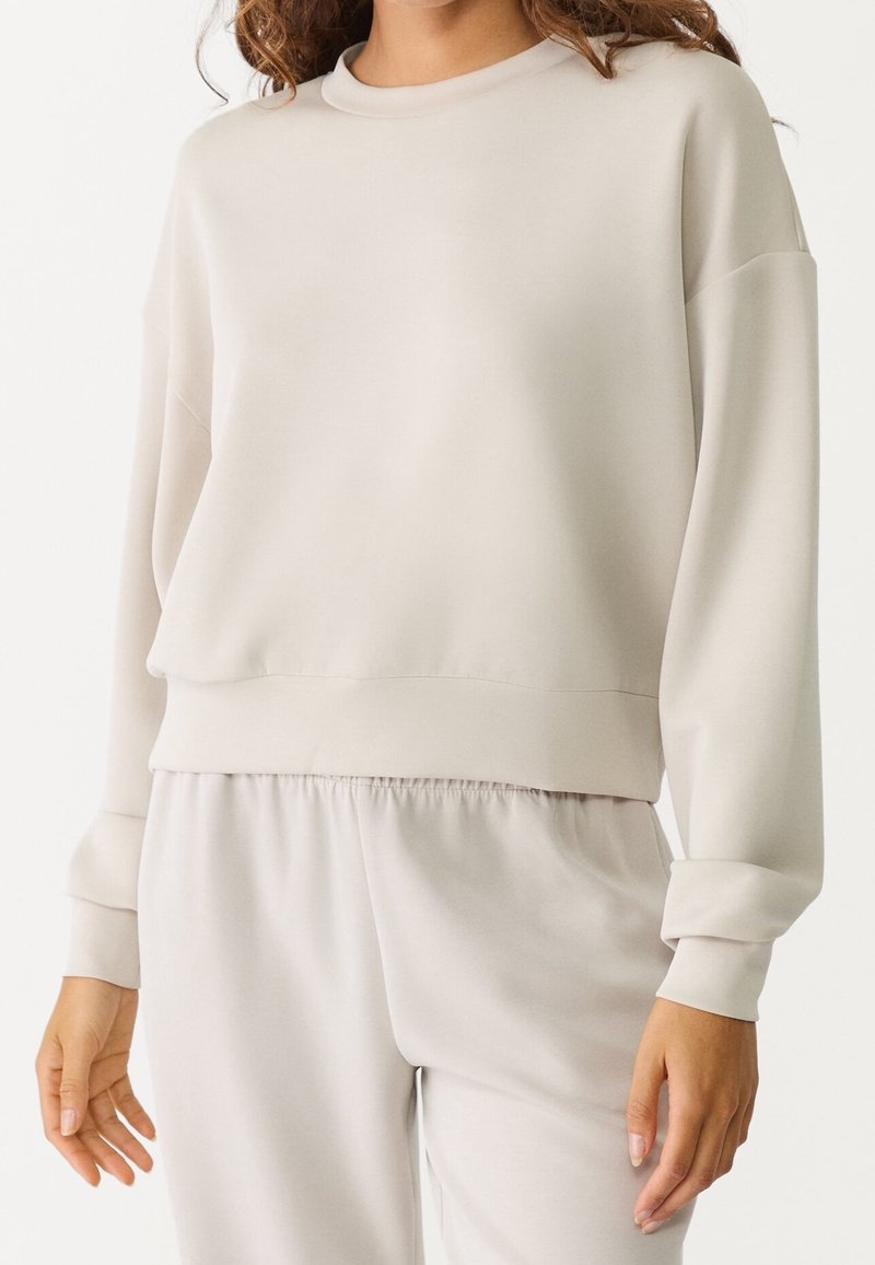 Femme portant un sweat-shirt à manches longues beige clair et un pantalon assorti avec taille élastique, debout devant un fond uni.