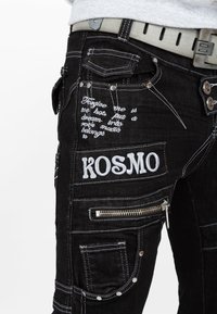 I pantaloni cargo in denim nero presentano testo ricamato, cerniere e molteplici tasche. Gli elementi distintivi includono cuciture bianche e una cintura con accessori metallici.