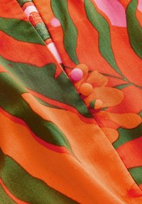 Orange tyg med stora blommönster i nyanser av grönt och rosa, med en slät textur och små, färgglada tygpuffar.