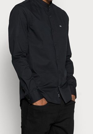 Homme portant une chemise noire boutonnée avec un petit logo blanc CK sur la poitrine et un pantalon noir, les mains dans les poches.