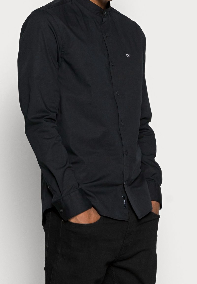 Homme portant une chemise noire boutonnée avec un petit logo blanc CK sur la poitrine et un pantalon noir, les mains dans les poches.
