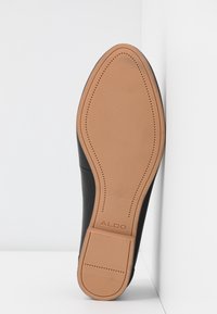 Chaussure en cuir noir avec semelle en caoutchouc tan lisse et petit talon, placée verticalement en angle contre un fond blanc d'angle, portant le nom de la marque "ALDO".