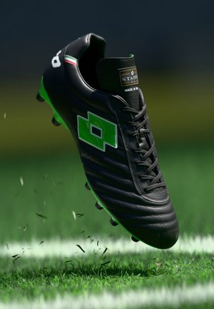 Black and green soccer cleat with laces, a green logo patch, and spikes kicking up grass on a marked soccer field.