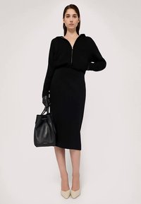 Svart zip-up hoodie kombinerad med en figurnära svart midikjol. Modellen håller en svart läderbucketväska och bär krämfärgade spetsiga klackskor.