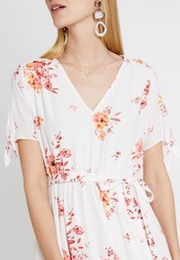 Vit klänning med blommönster i nyanser av rosa och orange, med V-ringning, korta ärmar och knytning i midjan. Enkla smycken i guld.