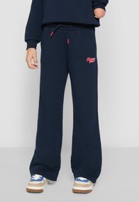 Jūras spēku sporta bikses no mīksta materiāla, ar auklu jostu, platām kājām un sarkanu izšūtu logotipu uz kreisās puses.