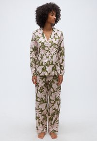 Γυναίκα που στέκεται ξυπόλητη, φοράει πυτζάμες σε πράσινο ελιάς με μεγάλα ανοιχτό ροζ ανθικά σχέδια, με πουκάμισο με κουμπιά και χαλαρά παντελόνια, κοιτάζοντας προς το πλάι.