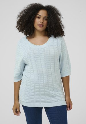 Vrouw met krullend haar, gekleed in een lichtblauwe gebreide trui met textuur, een scallopvormige halslijn en ellebooglange mouwen, gecombineerd met een blauwe spijkerbroek.