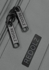 Grau strukturiert Tasche mit zwei schwarzen Reißverschlüssen, die mit "REDOLZ" beschriftet sind, und einem schwarzen Akzentpatch, das denselben Namen zeigt.