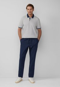 Herren-Polohemd in Weiß mit navyblauem Muster, kombiniert mit navyblauen Jogginghosen mit Kordelzug. Lässiges Schuhwerk mit weißem Obermaterial und beiger Sohle.