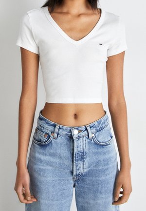 Kobieta ubrana w biały, krótki top z krótkim rękawem i dekoltem w serek oraz jasnoniebieskie dżinsy z wysokim stanem, stojąca na gładkim tle.