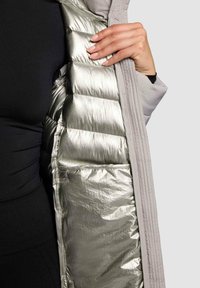 Gewatteerde jas met een zilveren binnenzijde en een grijze buitenkant; voorzien van doorgestikte details. Rits aan de voorkant en zichtbare zijzak.