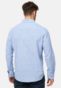 Chemise à manches longues bleu clair avec un col boutonné, en tissu texturé, et un ourlet arrondi. Présente des poignets boutonnés et un motif subtil.