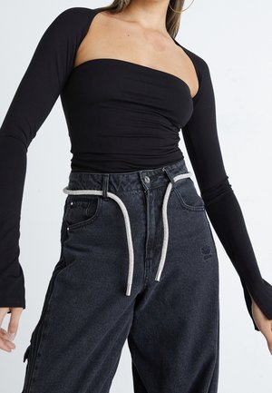Kobieta w czarnej bluzce z długim rękawem, odsłaniającej ramiona, oraz luźnych czarnych dżinsach z białym pasem sznurkowym na jednolitym tle.