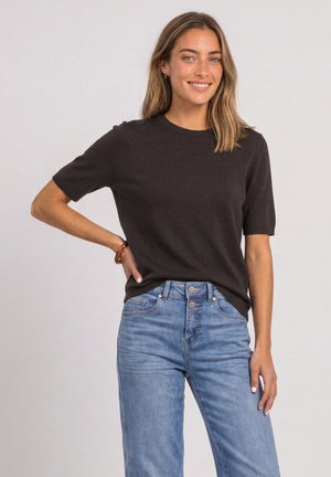 Femme souriante aux cheveux blonds mi-longs, portant un t-shirt noir à manches courtes et un jean bleu clair taille haute, debout avec une main sur la hanche.