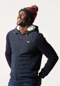 Marineblauwe puffer vest met een ritssluiting aan de voorkant en oranje accenten, gecombineerd met een donkere muts met een rood en zwart patroon en een pompondetail.