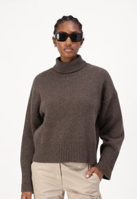 Καφέ, χοντρό πλεκτό πουλόβερ με ψηλό λαιμό και ριγωτές μανσέτες και στρίφωμα. Το μοντέλο φοράει μαύρα ορθογώνια γυαλιά ηλίου. Ανοιχτόχρωμο, ουδέτερο παντελόνι.