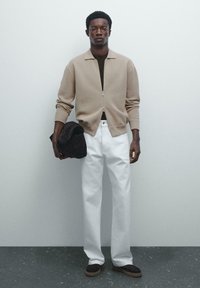 Un homme se tient contre un mur gris, portant une veste zippée beige, une chemise sombre, un pantalon blanc, des chaussures sombres, tenant une veste noire pliée dans une main.