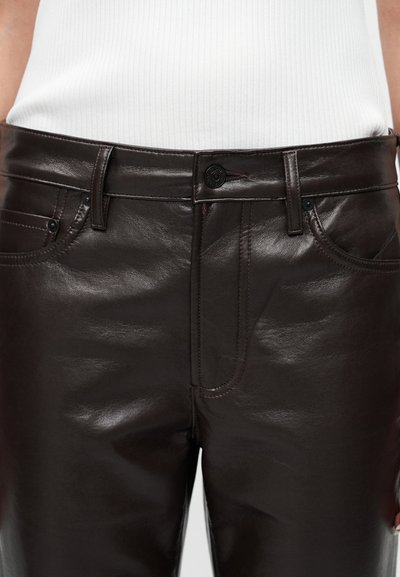 Κοντινό πλάνο σκούρων καφέ δερμάτινων παντελονιών με μπροστινές τσέπες, θηλιές για ζώνη και κλείσιμο με κουμπί, φορεμένα με λευκό ριπ τοπ.