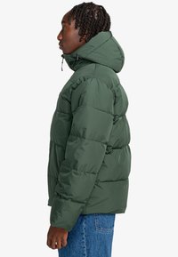 Giacca a vento verde con cappuccio, caratterizzata da sezioni trapuntate, un colletto alto e cordini regolabili. Indossata con jeans in denim blu.