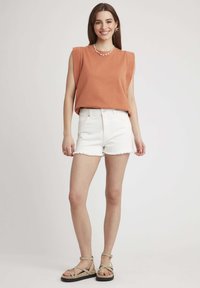 Top sans manches couleur rouille, short en denim blanc avec ourlet effiloché et sandales à lanières couleur beige. Tenue d'été simple et décontractée avec des détails minimaux.