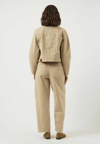 Žena s hnědými vlasy po ramena, v béžové džínové bundě a odpovídajících kalhotách s vysokým pasem, stojící zády k bílému pozadí.