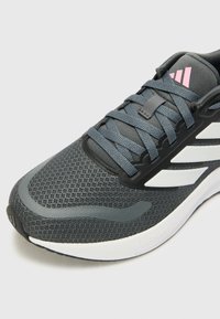 Chaussure de sport gris foncé avec un dessus en maille, lacets ton sur ton, semelle intermédiaire blanche et un accent de marque rose avec un design à trois rayures.