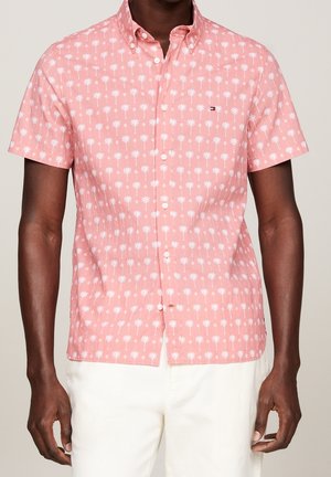 Man draagt een roze overhemd met korte mouwen en witte palmbomenprint en een witte broek tegen een neutrale achtergrond.